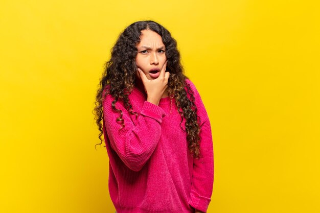 Jeune femme hispanique avec la bouche et les yeux grands ouverts et la main sur le menton se sentant désagréablement choquée de dire quoi ou wow