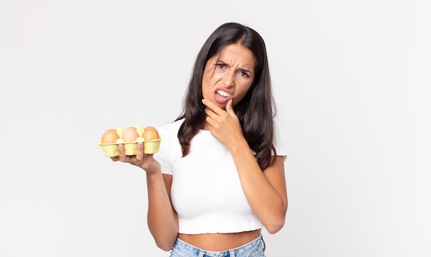 Jeune femme hispanique avec la bouche et les yeux grands ouverts et la main sur le menton et tenant une boîte à œufs