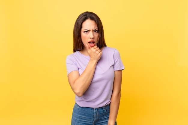 Jeune femme hispanique avec la bouche et les yeux grands ouverts et la main sur le menton