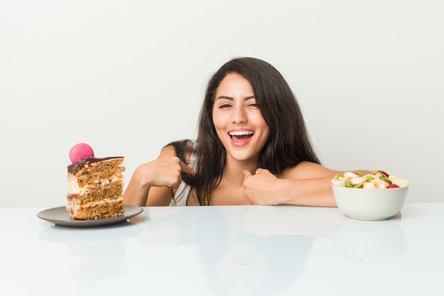 Jeune femme hispanique choisissant entre un gâteau ou un fruit surpris se montrant du doigt, souriant largement.