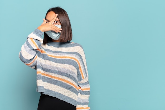 Jeune femme hispanique à choqué, effrayé ou terrifié, couvrant le visage avec la main et furtivement entre les doigts