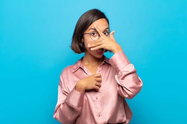 Jeune femme hispanique à choqué, effrayé ou terrifié, couvrant le visage avec la main et furtivement entre les doigts