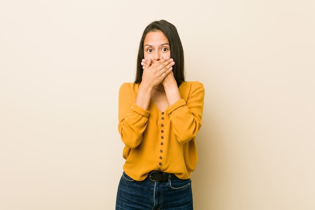 Jeune femme hispanique contre un mur beige choqué couvrant la bouche avec les mains.