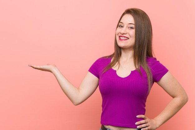 Jeune femme hispanique contre un mur rose montrant un espace copie sur une paume et tenant une autre main à la taille.