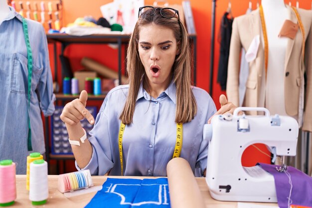 Jeune femme hispanique couturière designer à la salle d'atelier pointant vers le bas avec les doigts montrant la publicité, le visage surpris et la bouche ouverte