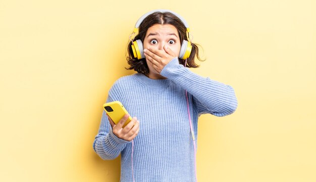 Jeune femme hispanique couvrant la bouche avec les mains avec un choc. casque et concept de smartphone