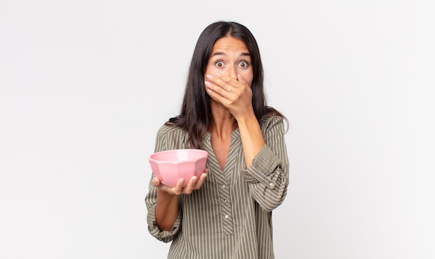 Jeune femme hispanique couvrant la bouche avec les mains avec un choc et tenant un bol ou un pot vide