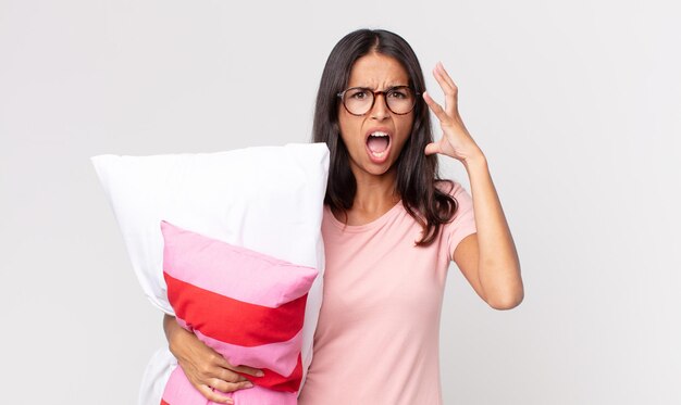 Jeune femme hispanique criant avec les mains en l'air portant un pyjama et tenant un oreiller