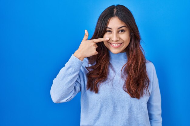 Jeune femme hispanique debout sur fond bleu pointant avec le doigt de la main vers le visage et le nez souriant concept de beauté joyeuse