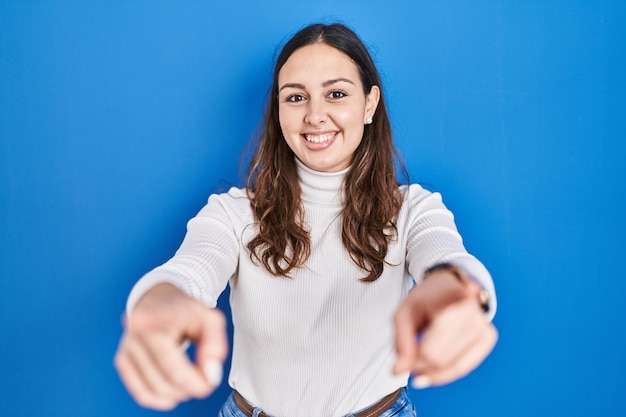 Photo jeune femme hispanique debout sur fond bleu pointant vers vous et la caméra avec des doigts souriants positifs et joyeux