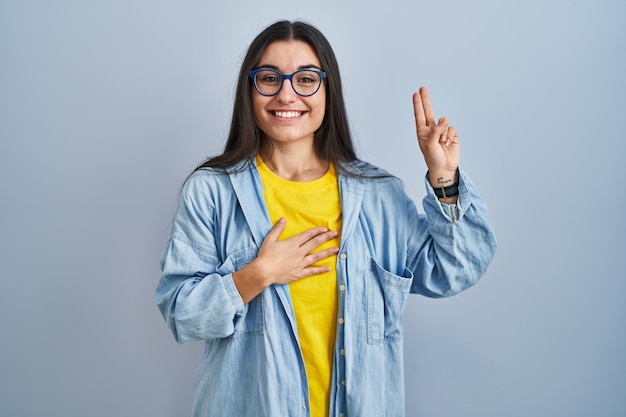 Jeune femme hispanique debout sur fond bleu souriant jurant avec la main sur la poitrine et les doigts vers le haut, faisant un serment de promesse de fidélité