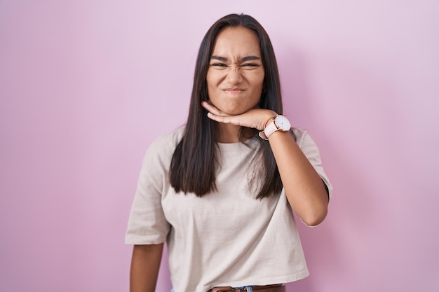 Jeune femme hispanique debout sur fond rose coupant la gorge avec la main alors que le couteau menace l'agression avec une violence furieuse