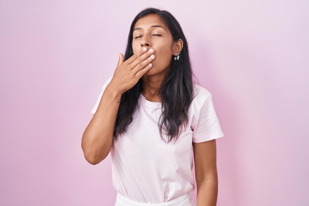 Jeune femme hispanique debout sur fond rose ennuyé bâillant fatigué couvrant la bouche avec la main agitée et somnolence