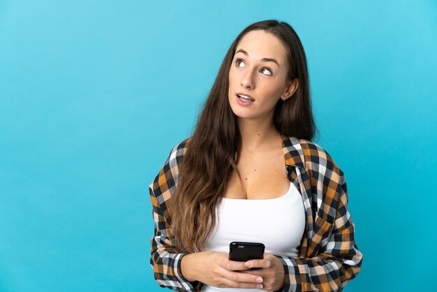 Jeune femme hispanique sur fond bleu isolé à l'aide d'un téléphone portable et levant