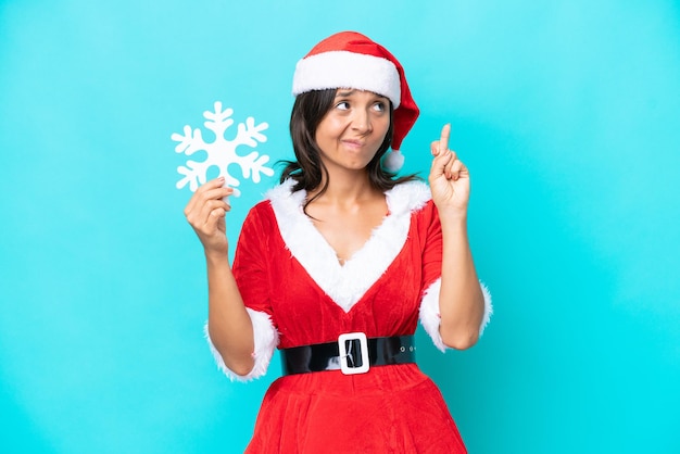 Jeune femme hispanique habillée en maman noel tenant un flocon de neige isolé sur fond bleu avec les doigts croisés et souhaitant le meilleur