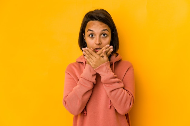 Jeune femme hispanique isolée sur bouche couvrant jaune choqué avec les mains