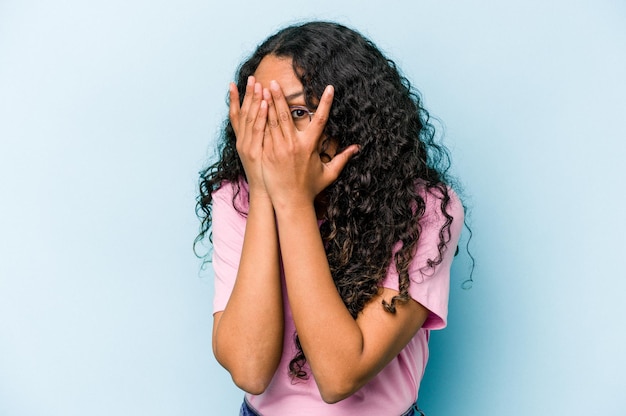 Jeune femme hispanique isolée sur fond bleu clignote à travers les doigts effrayée et nerveuse