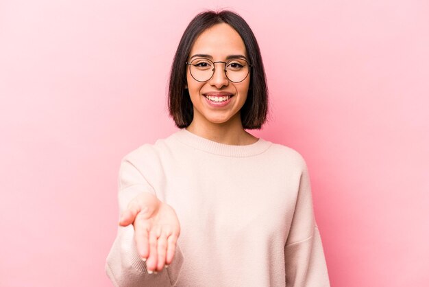Jeune femme hispanique isolée sur fond rose, tendant la main à la caméra en geste de salutation