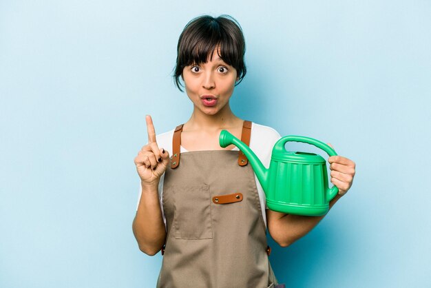 Jeune femme hispanique de jardinier tenant un arrosoir isolé sur fond bleu ayant une excellente idée de concept de créativité