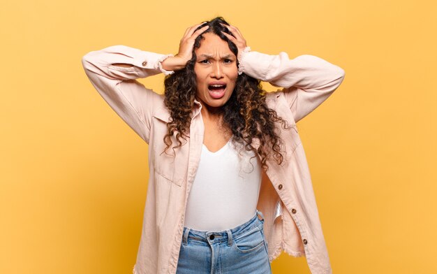 Jeune femme hispanique levant les mains à la tête, bouche bée, se sentant extrêmement chanceux, surpris, excité et heureux