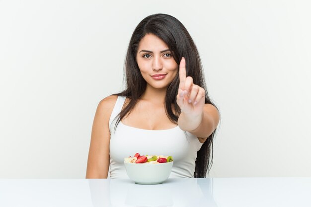 Jeune femme hispanique mangeant un bol de fruits montrant le numéro un avec le doigt.