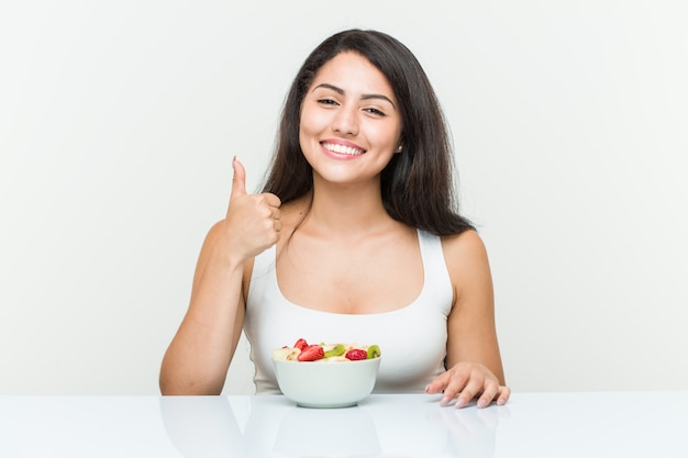 Jeune femme hispanique mangeant un bol de fruits souriant et levant le pouce vers le haut