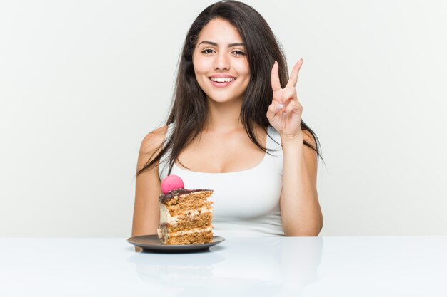 Jeune femme hispanique mangeant un gâteau montrant le numéro deux avec les doigts.