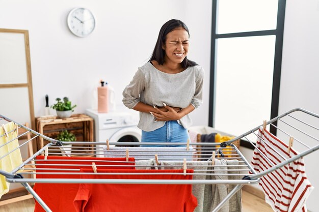 Jeune femme hispanique mettant du linge frais sur une corde à linge avec la main sur le ventre parce que l'indigestion, la maladie douloureuse se sent mal. notion de mal.