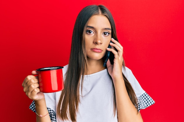 Jeune femme hispanique parlant sur le smartphone et buvant une tasse de café détendue avec une expression sérieuse sur le visage. simple et naturel en regardant la caméra.