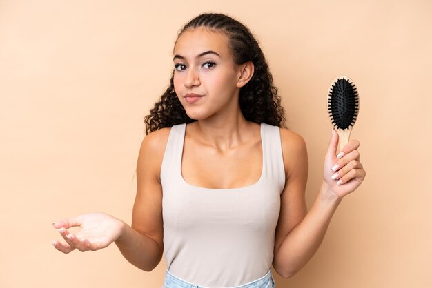 Jeune femme hispanique avec un peigne à cheveux isolé sur fond beige faisant un geste de doute tout en soulevant les épaules