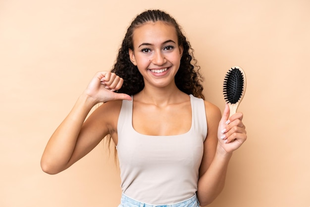 Jeune femme hispanique avec peigne à cheveux isolé sur fond beige fier et satisfait de soi