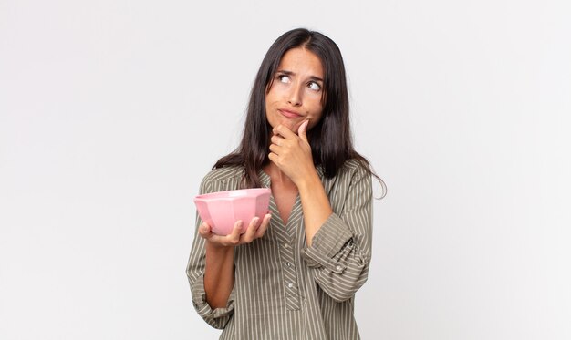 Jeune femme hispanique pensant, se sentant dubitative et confuse et tenant un bol ou un pot vide