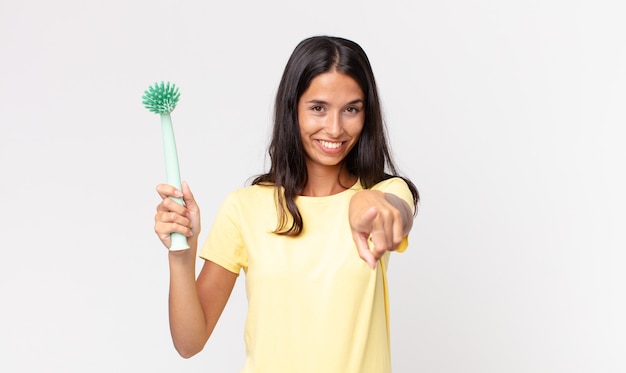 Jeune femme hispanique pointant vers la caméra vous choisissant et tenant une brosse à vaisselle