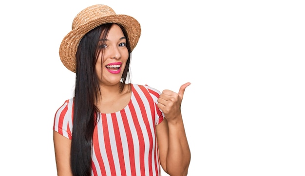 Jeune femme hispanique portant un chapeau d'été souriant avec un visage heureux regardant et pointant vers le côté avec le pouce vers le haut.