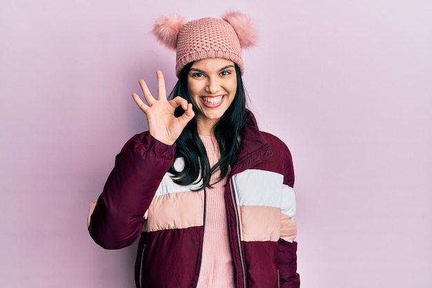 Jeune femme hispanique portant un pull d'hiver en laine et une casquette souriante positive faisant signe ok avec la main et les doigts. expression réussie.