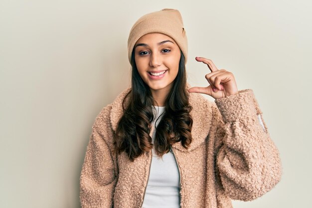 Jeune femme hispanique portant un pull de laine et un chapeau d'hiver souriante et confiante faisant des gestes avec la main faisant un signe de petite taille avec les doigts en regardant et le concept de mesure de la caméra