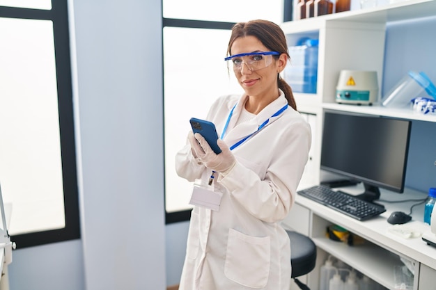Une jeune femme hispanique portant un uniforme de scientifique utilisant un smartphone au laboratoire