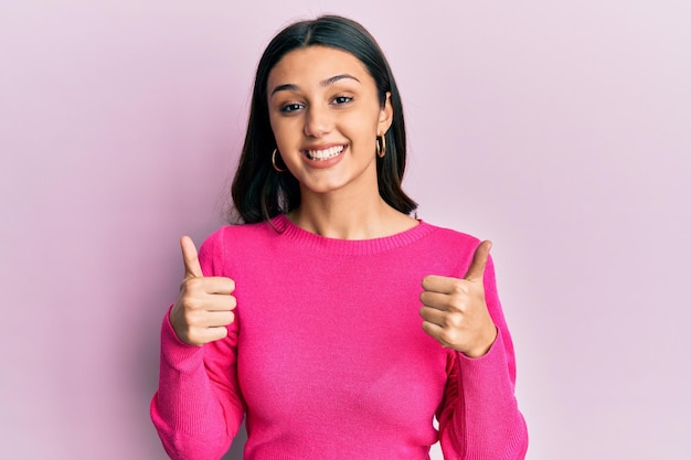 Jeune femme hispanique portant des vêtements décontractés signe de succès faisant un geste positif avec les pouces de la main en l'air souriant et heureux expression joyeuse et geste de gagnant