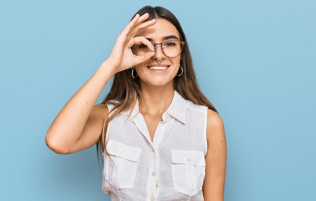 Jeune femme hispanique portant des vêtements décontractés souriant heureux faisant signe ok avec la main sur les yeux regardant à travers les doigts