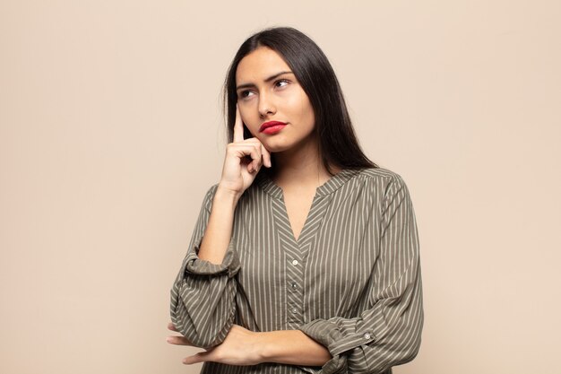 Jeune femme hispanique avec un regard concentré, se demandant avec une expression douteuse, regardant vers le haut et sur le côté