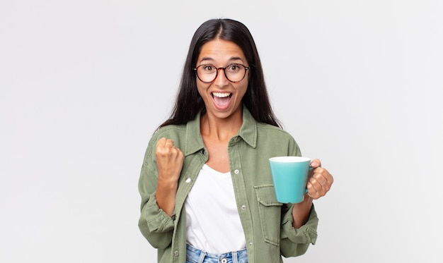 Jeune femme hispanique se sentant choquée, riant et célébrant le succès et tenant une tasse de café