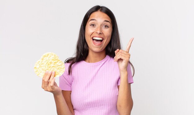 Jeune femme hispanique se sentant comme un génie heureux et excité après avoir réalisé une idée et tenant un biscuit au riz. concept de régime