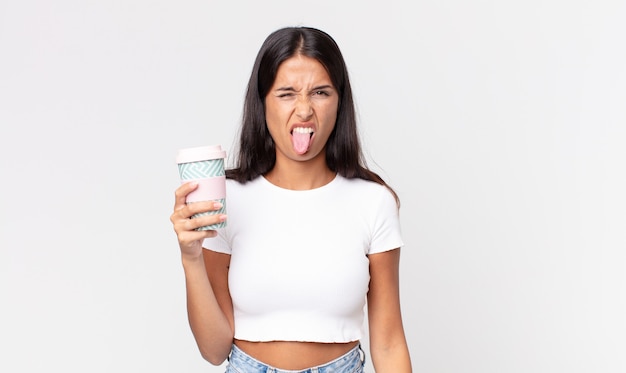 Jeune femme hispanique se sentant dégoûtée et irritée et tirant la langue et tenant un récipient de café à emporter