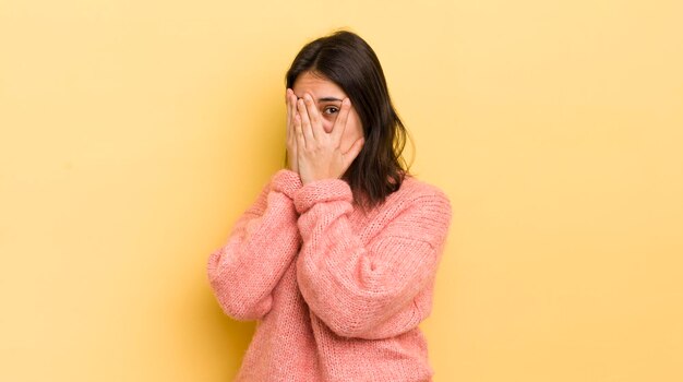 Jeune femme hispanique se sentant effrayée ou gênée de jeter un coup d'œil ou d'espionner avec les yeux à moitié couverts de mains