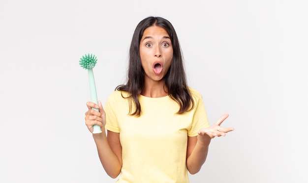 Jeune femme hispanique se sentant extrêmement choquée et surprise et tenant une brosse à vaisselle