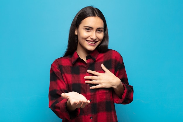 Jeune femme hispanique se sentant heureuse et amoureuse, souriante d'une main à côté du cœur et de l'autre tendue vers l'avant