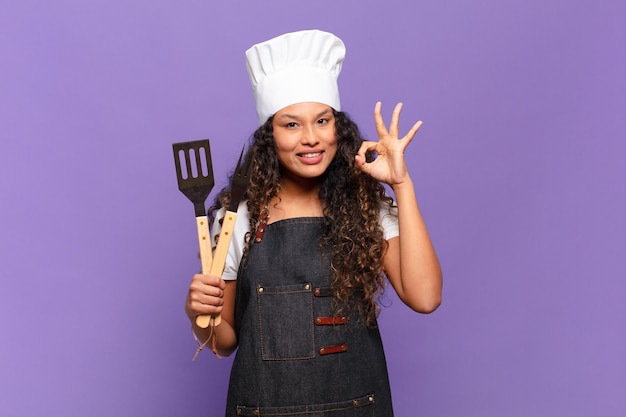 Photo jeune femme hispanique se sentant heureuse, détendue et satisfaite, montrant son approbation avec un geste correct, souriant. concept de chef de barbecue