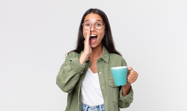 Jeune femme hispanique se sentant heureuse, donnant un grand cri avec les mains à côté de la bouche et tenant une tasse de café