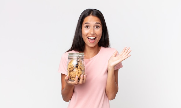 Jeune femme hispanique se sentant heureuse et étonnée de quelque chose d'incroyable et tenant une bouteille en verre de biscuits