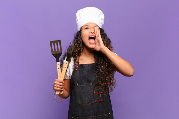 Jeune femme hispanique se sentant heureuse, excitée et positive, donnant un grand cri avec les mains à côté de la bouche, appelant. concept de chef de barbecue